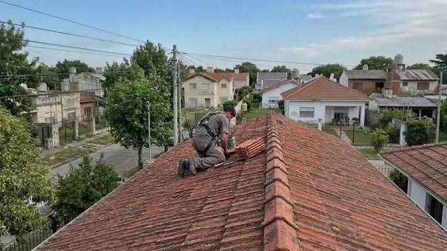 Techista con arnés recolocando tejas cerámicas en el techo a dos aguas de una casa de barrio